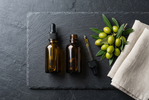A full and a nearly empty amber glass dropper bottle of liquid vitamin D on slate, beside green olives and a linen cloth.