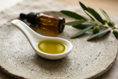 A white ceramic spoon holding a small pool of golden-green olive oil, with olive leaves and an amber dropper bottle softly out of focus behind it.