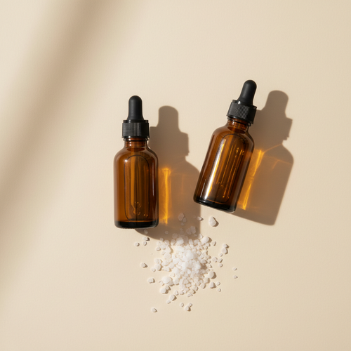 Two amber glass dropper bottles side by side on a cream background with a small scattering of white mineral granules between them