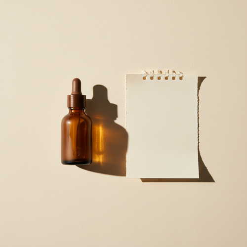 An amber glass dropper bottle beside a blank curled calendar page on a cream background, lit to cast a long shadow
