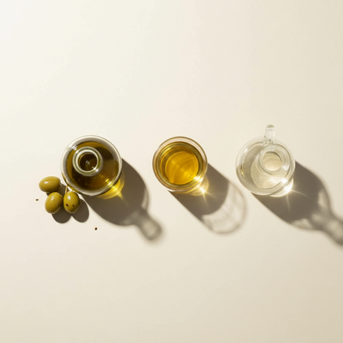 Three small glass vessels of oil in different shades, from golden green to pale clear, arranged in a row beside fresh olives on a cream background